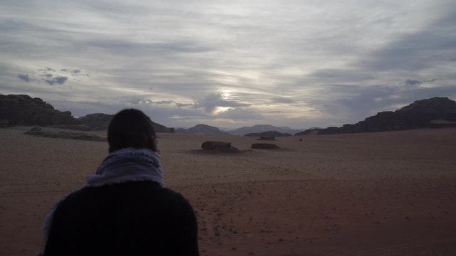 man standing on the edge of a cliff in the desert of wadi rum and covering his face with the arabic scarf