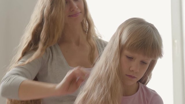 Close-up hands of young woman combing hair of her blond daughter. Concept of motherhood, childhood, one parent. Leisure indoors. Happy family