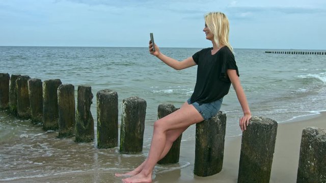 Slow motion zoom in young woman takes a selfie at the beach seated on a fence post