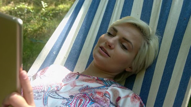 Close-up static shot of a middle aged blonde lady laying down on a hammock reading on a tablet