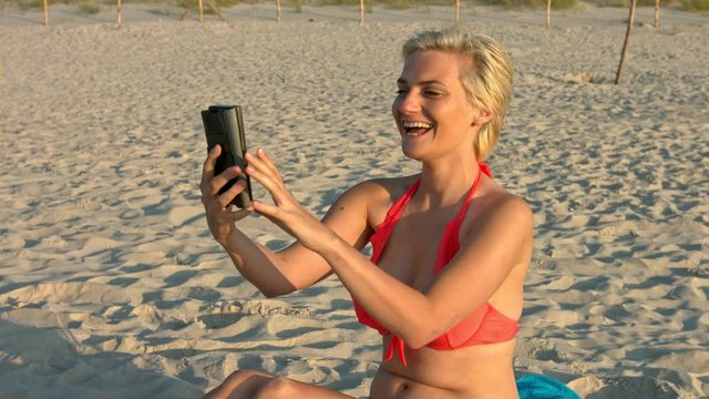 Happy short blonde hair woman video calling through her mobile phone device at a beach wearing bikini swimwear while sitting on white sand during sunny day time