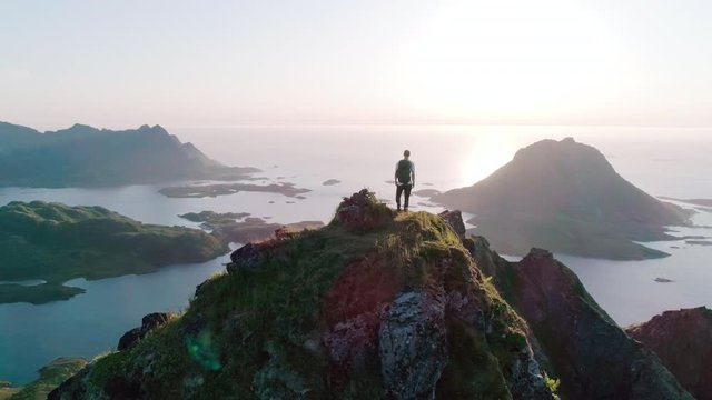 4K drone shot of Norwegian climber standing on the top of a tall mountain looking at and admiring the panorama sunset background view of Lofoten and Vesterålen in Norway.