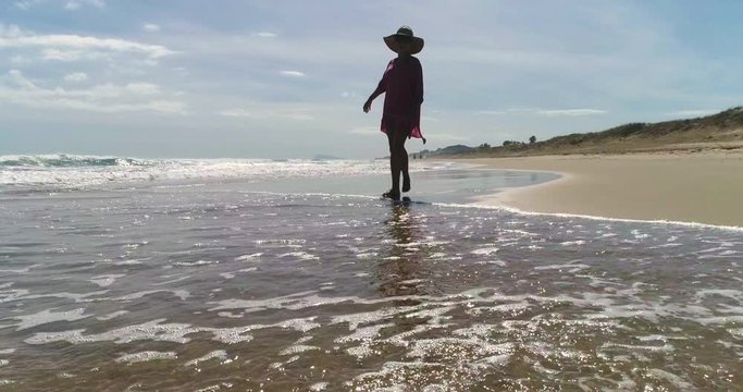 Dron vuela ras del suelo siguiendo a una mujer que pasea por la orilla