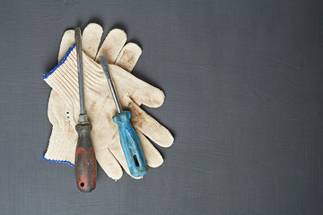 Pair of dirty textile protective gloves for repair or other hard works near old messy screwdrivers with plastic handles lies on dark scratched concrete table in workroom. Copy space. Top view