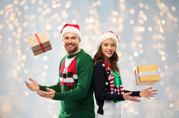 people and holidays concept - happy couple in santa hats with christmas gifts at ugly sweater party over festive lights background