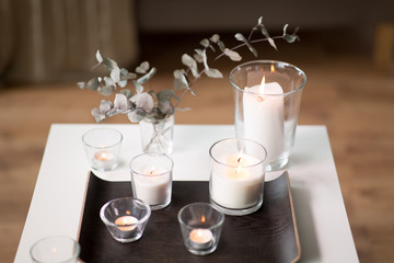 decoration, hygge and cosiness concept - burning white fragrance candles on tray and branches of eucalyptus populus on table at cozy home