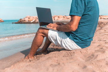 Adult male of mediterranean race using laptop while sitting on the sea beach. Freedom and travel concept