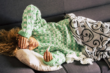 Fototapeta na wymiar alone woman at home lying down on the sofa looking at the thermometer with high fever themperature - sick lady with curly hair using tissues and blanket for seasonal winter cold illness