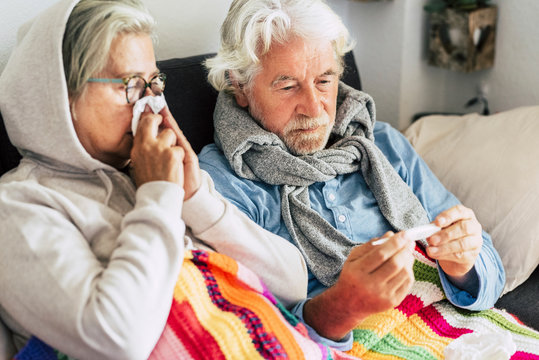 couple of sicks seniors and mature people sitting at the sofa with fever looking at the thermometer with high temperature - two elderly woman and man sick with illness for seasonal cold