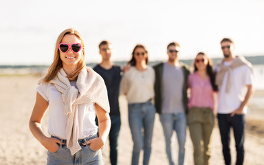 Obraz premium friendship, valentine's day and people concept - happy woman in heart-shaped sunglasses with group of friends on beach in summer