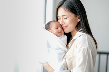 White shirt Asian mother is kissing her newborn baby in bedroom in front of glass windows with white curtain to show love and family bonding.