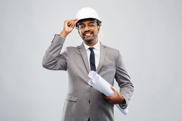 architecture, construction business and people concept - smiling indian male architect in helmet with blueprints over grey background