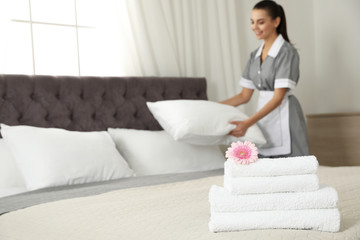 Stack of clean towels with flower on bed and blurred chambermaid in hotel room, closeup. Space for text