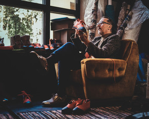 Mature shoe designer is sitting on the armchair at his workshop and holding a shoe from his last collection.