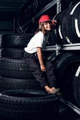 Happy smiling girl in cap is sitting on tyres at dark tyres warehouse.