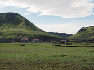 Obraz premium Hvanngil campsite path in lava field and green valley, small houses of hvanngil hut. volcanic mountains volcanic landscape with blue sky, Laugavegur Trail between Emstrur-Botnar and Alftavatn, central