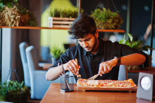 Confident Young Indian Man In Black Shirt Sitting At Pizzeria With Pizza.