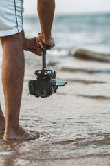 part of the body of a young man with a camera attached to a steadicam, a videographer shoots video walking barefoot along the sandy seashore, a concept of hobby and relaxation