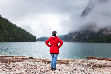 Fototapeta na wymiar Women with red rain jacket standing all alone on a rocky mountain lake bank on a cloudy misty rainy autumn day and enjoying the peacful nature view after a hike.