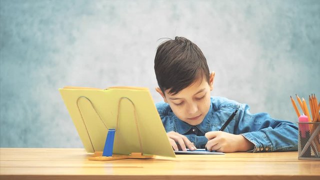 Young concentrated school kid in blue jeans t-shirt writing, copying the text from book on the bookholder.