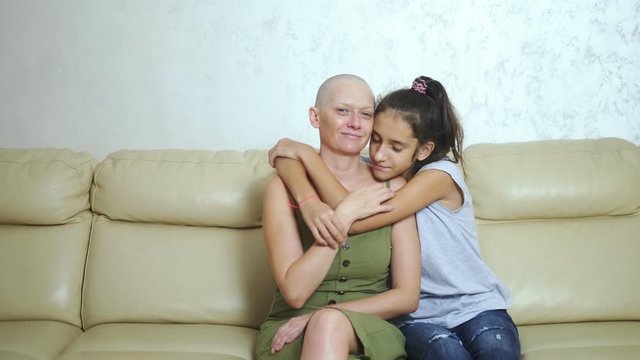 bald exhausted mother after oncology chemotherapy hugs her teenager daughter while sitting on the couch