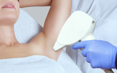 The cosmetologist does the laser hair removal procedure in the armpit zone, to a young woman in a...