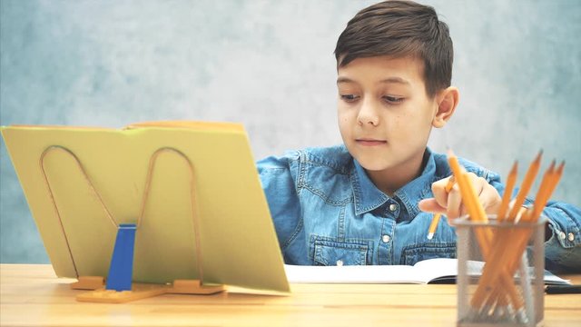 Young concentrated school kid in blue jeans t-shirt writing, copying the text from book on the bookholder, reaching the ruler, pencil and underlining.