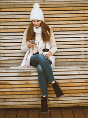 Beautiful young girl drinking coffee, tea from plastic mug in autumn, winter and talking on mobile phone. Woman with long hair sitting on bench in autumn or winter, basking in hot drink, copy space