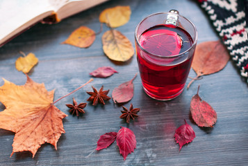 An open book, a warm scarf and a beautiful red tea with mulled wine lie on a gray shabby  table among yellow autumn leaves and anise stars