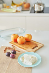 ingredients for baked apples: apples, plums, cottage cheese and cinnamon on a table in a white kitchen