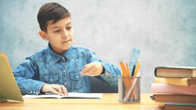 Young concentrated school kid in blue jeans t-shirt appearing on the gray background, sitting down, writing with a highlighter.