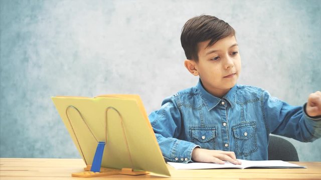 Young concentrated school kid in blue jeans t-shirt appearing on the gray background, sitting down, opening the copybook, writing with a highlighter.