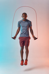 Staying fit. Full length of young african man in sport clothing skipping rope while exercising in studio against colorful background - obrazy, fototapety, plakaty