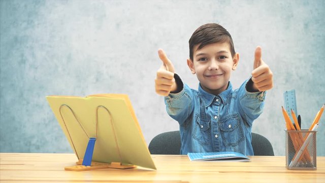 Young clever school kid in blue jeans t-shirt was writing, finished his homework, closed the copybook and now satisfied with the result, pointing thumbs up.