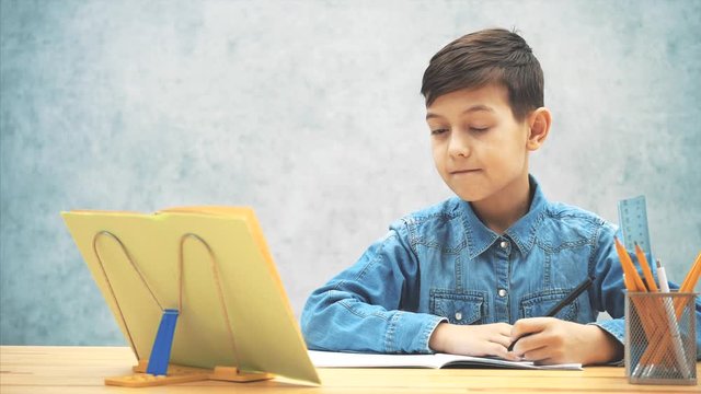 Young concentrated school kid in blue jeans t-shirt writing, copying the text from book on the bookholder. His sister joining him, looking how he is writing. He fends her off.