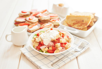 fried zucchini with tomatoes and sour cream sauce; salad with cheese, ham, toast and herbs on a white wooden table
