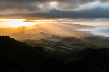 Stunning sunset view of the volcanic landscape on the island of São Miguel in the Azores archipelago. Volcanic cones are illuminated by sun rays under a dramatic cloudy sky with the Atlantic ocean.