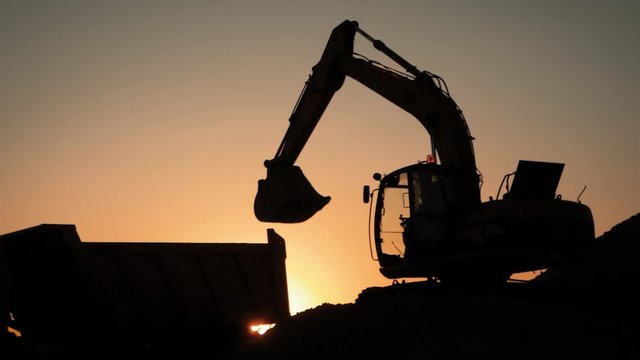 Silhouette of an excavator that loads sand into a truck at sunset. Concept construction and heavy industry, machine will be used in heavy industry business. Slow motion footage.