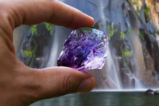 Hand Holding Beautiful Purple Crystal With Defocused Waterfall In The Background