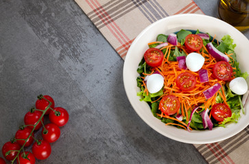 green leaf salad with carrot, tomato, onion and cheese on gray table with tomatoes and glass of olive oil top view