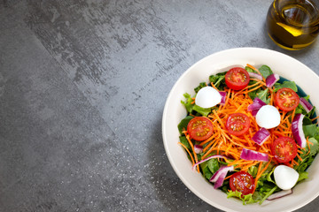 green leaf salad with carrot, tomato, onion and cheese on gray table with salt and glass of olive oil top view