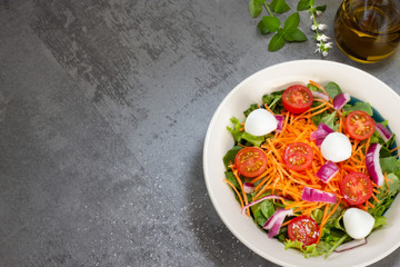 green leaf salad with carrot, tomato, onion and cheese on gray table with salt, glass of olive oil and basil leaves top view