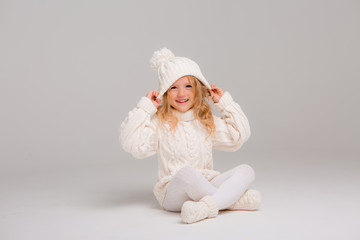 Winter clothes. Portrait of a little curly-haired girl in a knitted white winter hat. little blonde girl in white knitted hat and sweater smiling light background isolate, space for text