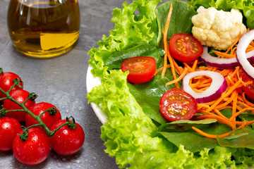 green leaf salad with carrot, tomato, onion, cauliflower with glass of olive oil in the background on gray surface
