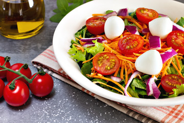 close up green leafy salad with carrot, tomato, onion and cheese near tomatoes on gray table with olive oil and basil leaves in the background