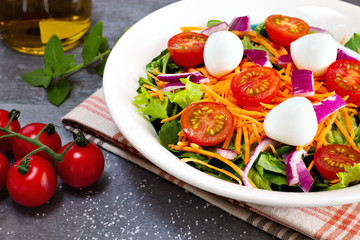 close up green leafy salad with carrot, tomato, onion and cheese near tomatoes on gray table with olive oil and basil leaves in the background