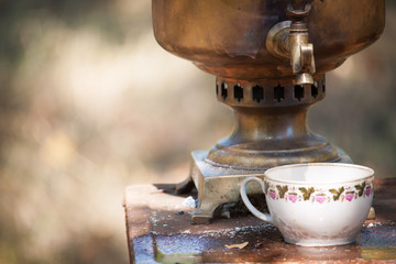 Vintage bronze samovar wood burning and cup on a rusty table outdoor, preparing water for tea brewing, selective focus