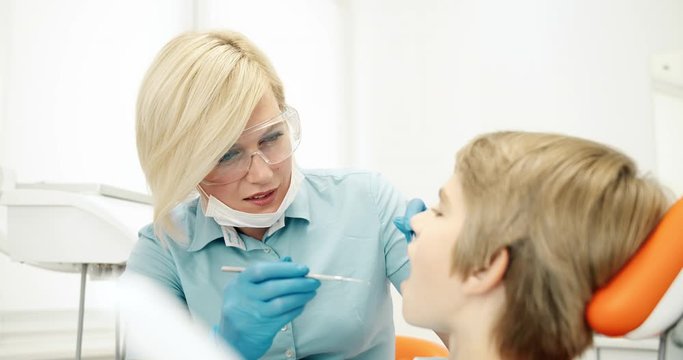 Professional lady dentist in protective eyeglasses checking boys teeth at regular oral cavity examination, healthcare and caries prevention