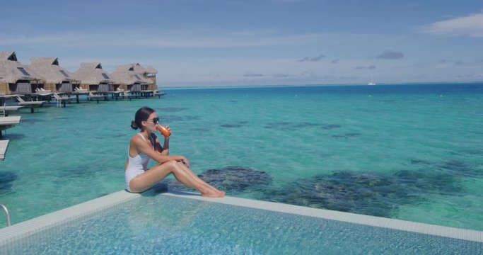 Healthy juice detox happy woman drinking carrot juicing smoothie glass on summer sunny day vacation background. Woman in bikini by luxury swimming pool in overwater bungalow. Elegant lady at resort.