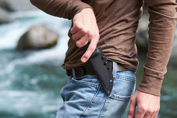 Man using the cross draw method to pull his hunting knife from it's kydex sheath while standing outdoors next to a mountain creek.